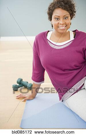 Stock Image - African woman sitting on yoga mat with dumbbells. Fotosearch