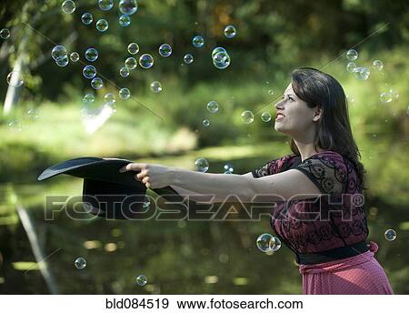 Stock Photo - Caucasian woman trying to catch bubbles in hat. Fotosearch