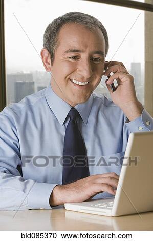 Stock Image - Hispanic businessman with laptop talking on cell phone. Fotosearch