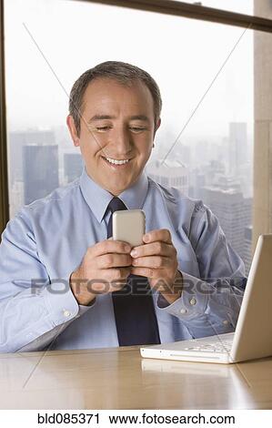 Stock Image - Hispanic businessman with laptop text messaging on cell phone. Fotosearch