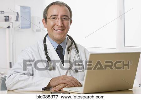 Stock Photo - Hispanic doctor sitting at desk with laptop. Fotosearch