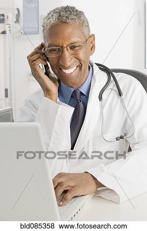 Stock Photo - Mixed race doctor sitting at desk talking on cell phone. Fotosearch