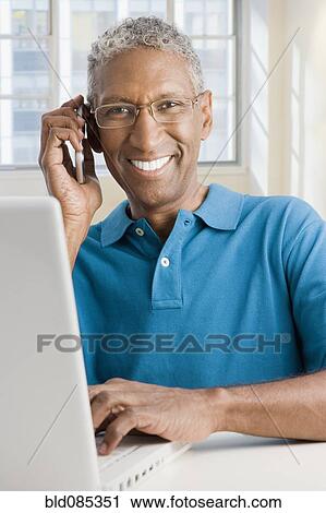 Stock Image - Mixed race man with laptop talking on cell phone. Fotosearch