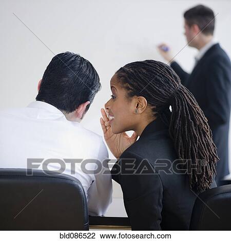 Stock Image - Businesswoman whispering to co-worker in meeting. Fotosearch