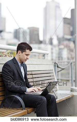 Stock Image - Mixed race businessman using laptop on city bench. Fotosearch