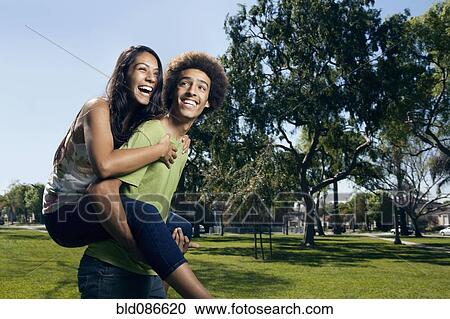 Stock Image - Teenager carrying girlfriend on his back in park. Fotosearch