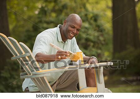 Stock Photo - African American man painting chair in backyard. Fotosearch