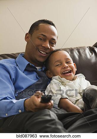 Stock Image - Father and son sitting on sofa watching television. Fotosearch