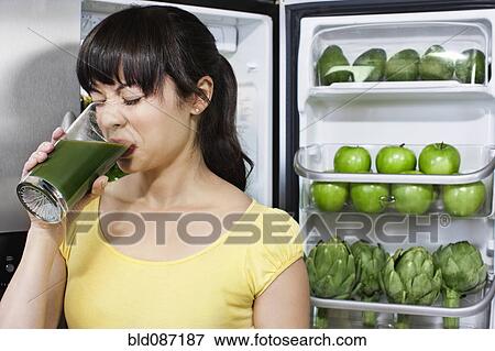Stock Photo - Grimacing mixed race woman drinking healthy drink near refrigerator. Fotosearch