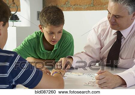 Stock Image - Hispanic father playing dominoes with sons. Fotosearch