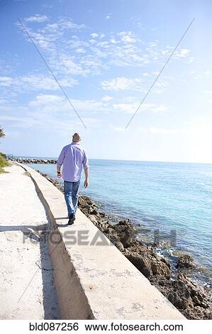 Stock Photograph - Hispanic man walking on wall near ocean. Fotosearch