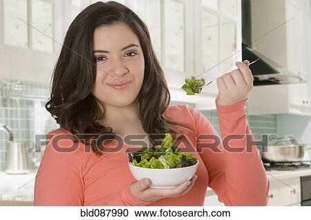 Stock Image - Hispanic teenage girl eating salad in kitchen. Fotosearch