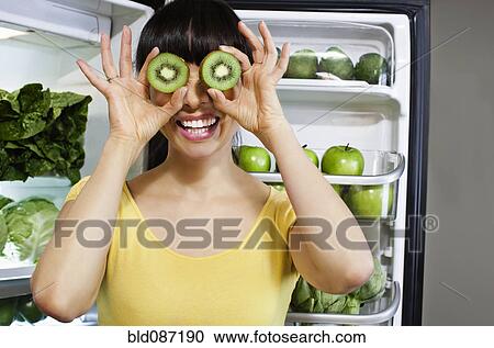 Stock Image - Mixed race woman covering eyes with kiwi slices near refrigerator. Fotosearch