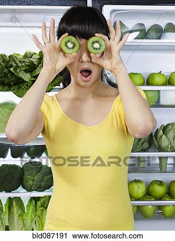 Stock Image - Mixed race woman covering eyes with kiwi slices near refrigerator. Fotosearch