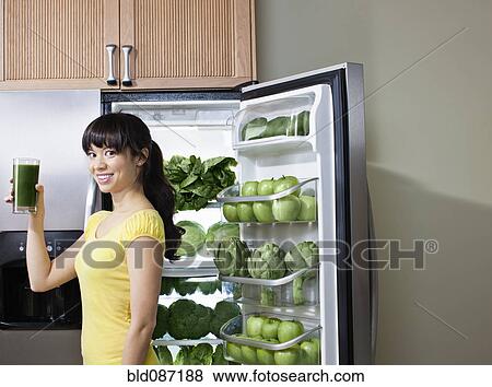 Stock Photo - Mixed race woman drinking healthy drink near refrigerator. Fotosearch