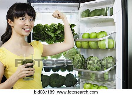 Stock Photo - Mixed race woman drinking healthy drink near refrigerator. Fotosearch