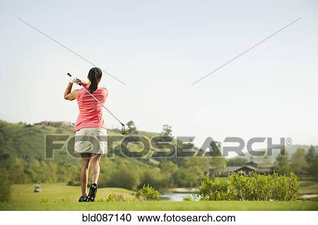 Stock Image - Mixed race woman swinging golf club on golf course. Fotosearch