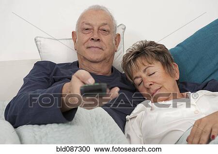 Stock Image - Senior Hispanic couple laying in bed watching television. Fotosearch