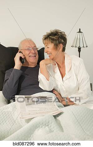 Stock Photograph - Senior Hispanic couple sitting in bed talking on telephone. Fotosearch