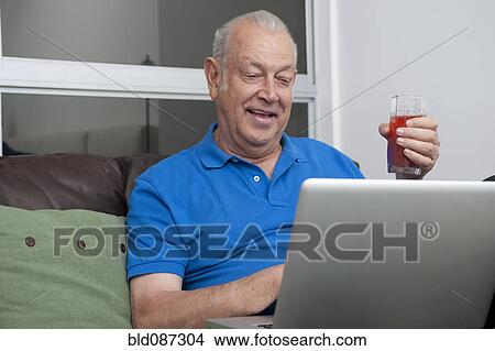 Picture - Senior Hispanic man holding drink and using laptop. Fotosearch