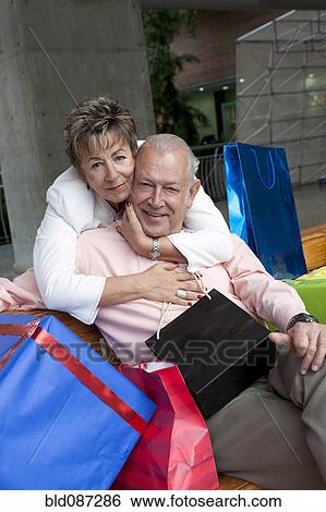 Stock Photograph - Senior Hispanic woman hugging husband surrounded by gifts. Fotosearch
