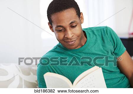 Stock Image - African American man reading book on bed. Fotosearch