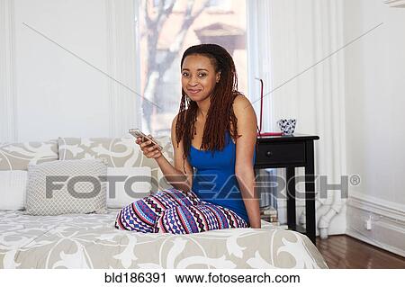 Stock Image - African American woman using cell phone on bed. Fotosearch