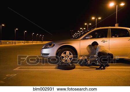 Stock Image - African businessman changing flat tire in parking lot. Fotosearch