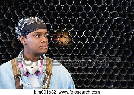 Stock Image - African female worker in front of stacks of metal pipes. Fotosearch