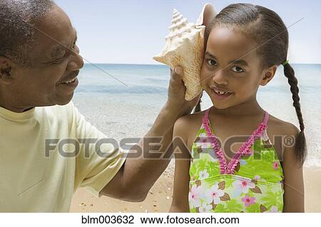 Stock Image - African grandfather holding conch shell to granddaughter's ear. Fotosearch