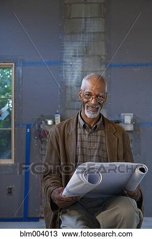 Stock Image - African man holding blueprints at construction site. Fotosearch