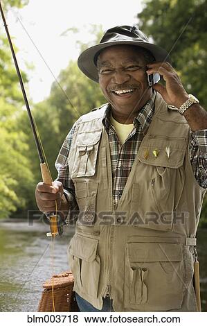 Stock Photo - African man in fishing vest using cell phone. Fotosearch