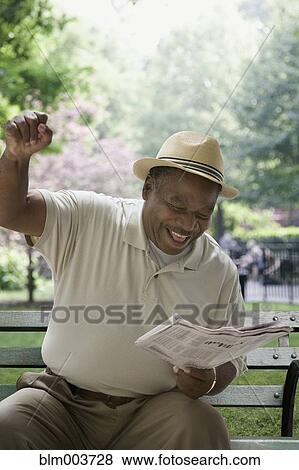Stock Photo - African man reading newspaper and gesturing on park bench. Fotosearch