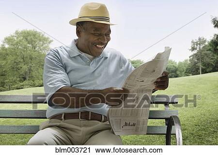 Stock Image - African man reading newspaper on park bench. Fotosearch