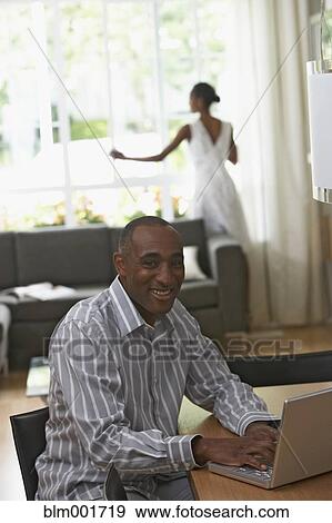 Stock Photo - African man typing on laptop with wife in background. Fotosearch