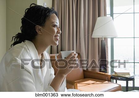 Stock Image - African woman drinking coffee in bedroom. Fotosearch