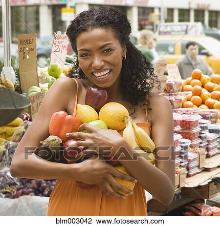 Stock Image - African woman holding armful of fruits and vegetables. Fotosearch