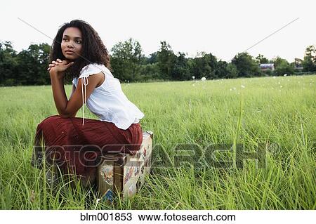 Stock Image - African woman sitting on suitcase in meadow. Fotosearch