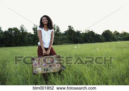 Stock Image - African woman standing with suitcase in meadow. Fotosearch