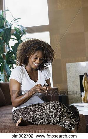 Stock Image - African woman text messaging on cell phone in living room. Fotosearch