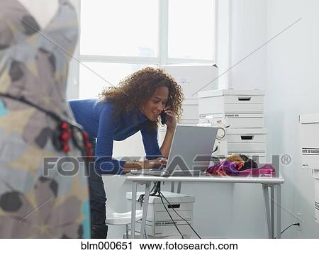 Stock Image - African woman typing on laptop and talking on cell phone. Fotosearch