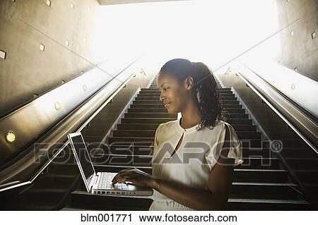 Stock Image - African woman typing on laptop in front of stairs. Fotosearch