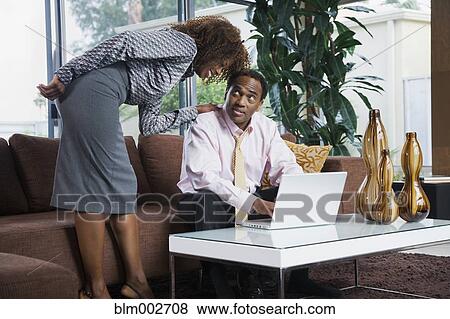 Stock Photo - African woman watching husband type on laptop in living room. Fotosearch