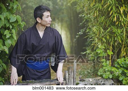 Stock Image - Asian man wearing traditional robe standing in forest. Fotosearch