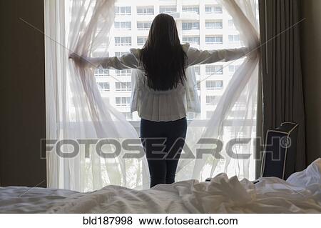 Stock Photo - Asian teenage girl opening curtains at hotel window. Fotosearch