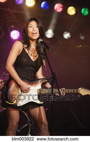 Stock Image - Asian woman singing and playing electric guitar onstage. Fotosearch