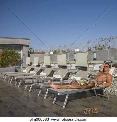 Stock Image - Bare chested man sunbathing in lounge chair poolside. Fotosearch