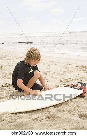 Stock Image - Boy waxing surfboard at beach. Fotosearch