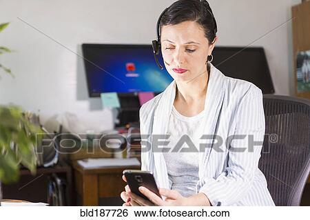 Businesswoman using cell phone and headset in office View Large Photo Image Stock Photograph - Businesswoman using cell phone and headset in office. Fotosearch