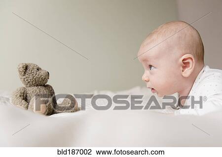 Stock Image - Caucasian baby staring at teddy bear on bed. Fotosearch